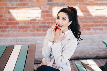 Portrait of surprised beautiful woman with dark pony tail sitting at cafe chatting with her best friend over smartphone gossiping having shocked look after hearing interesting and intriguing news