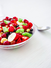 Fruit salad in glass plate with banana, kiwi and red berries on white rustic table.