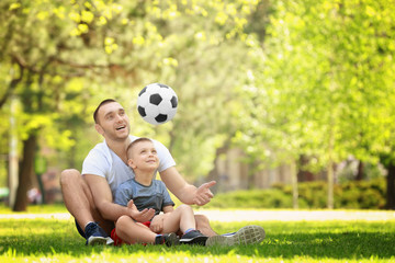 Naklejka premium Father and son playing with soccer ball while sitting on green grass in park