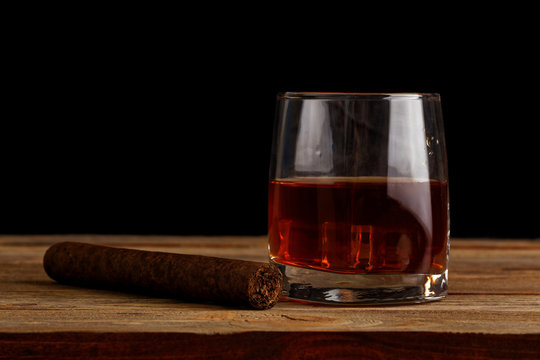Glass of whiskey with ice cubes and cigar on a wooden table against black background. Copyspace.