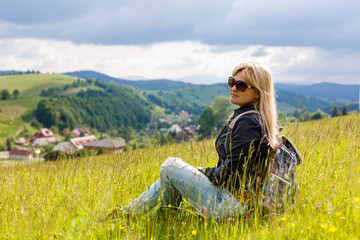 Naklejka na ściany i meble Smiling woman with backpack sitting in the grass with wild flowers on a hill and looking over the mountains, village in the valley and the rays of the sun breaking through the clouds at sunset