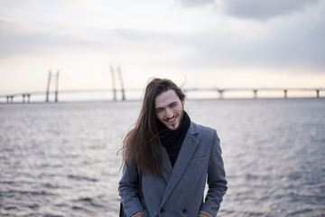 Outdoor portrait of fashionable handsome young bearded European man with long dark hair wearing elegant grey coat spending evening on promenade, smiling broadly, river and bridge in background