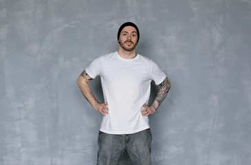 Attractive charismatic young dancer or sportsman with tattoos and earring wearing white t-shirt, black hat and grey jeans standing indoors, holding hands on his waist, ready for practise or training
