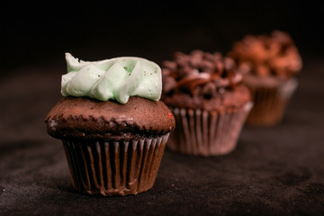 Three chocolate cupcakes garnished with chocolate chips and chocolate and mint frosting on lined up across the frame, on black background