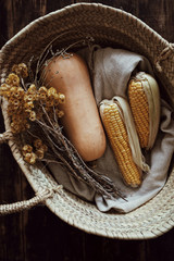 Top view on fresh healthy corn and pumpkin laying in straw rustic braided basket with linen textile and dried yellow flowers on dark wooden table background. Vertical atmospheric shot.