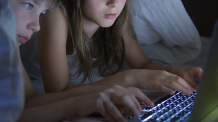 A boy and a girl are playing on the computer and lying on the bed under the blanket at night. - Powered by Adobe