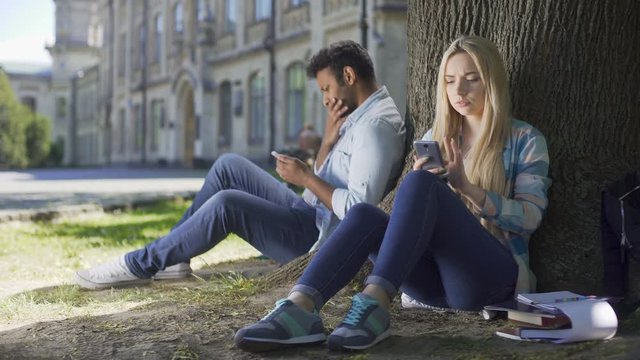 Male, female strangers sitting under tree, using cellphone, girl sighing heavily