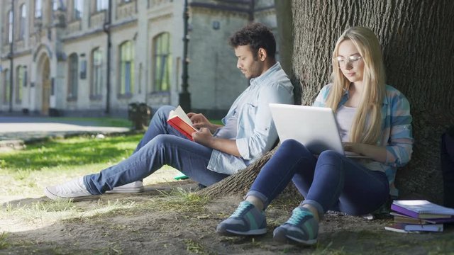 Male with book, female with laptop under tree looking at each other and smiling