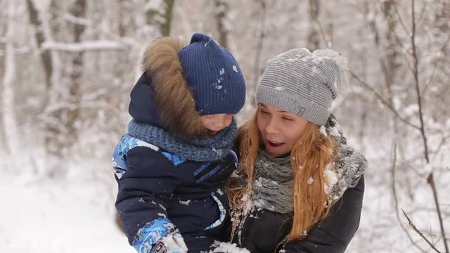Young mother holding a little boy during a snowfall in the woods on a walk. Mother and son playing with snow in winter forest.