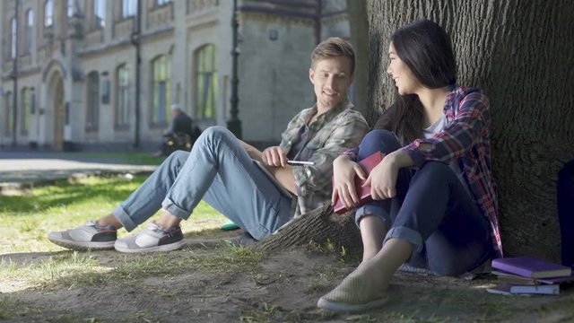 Multinational young man and woman sitting under tree, getting to know each other