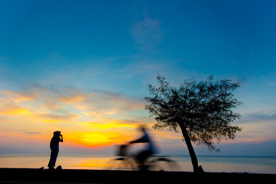 Silhouette of a young woman on the beach with motion blurred bicycle at sunrise. An image of a woman looking to the ocean. Travel and holiday concept.