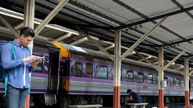 young asian man with backpack standing on platform at train station. backpacker or traveler using digital tablet while waiting for train. journey, trip, travel concept