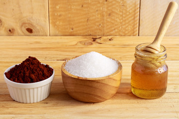wooden honey dipper in honey bottle and sugar in wooden bowl and cocoa in ceramic bowl on wooden table