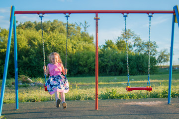 Portrait of a seven year old girl in a bright pink dress swinging on a swing in the park against a blue sky in the evening