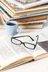 glasses lying on an open book next to the mug of coffee on background of stack of old books