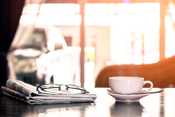Closeup cup of hot cappuccino coffee with newspaper and spectacles on the wood table with blur coffee shop background-warm tone.