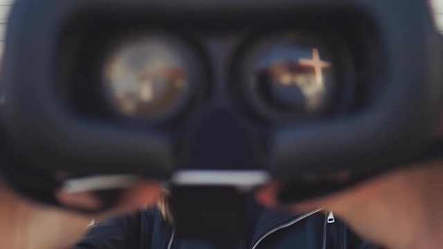 A young stylish blonde in a leather jacket near horizontal roller blinds brings virtual glasses to the camera.