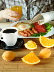 Breakfast and a woman who is reading a newspaper and is having a breakfast. Breakfast consisting of eggs, sausages, vegetables, fruit and drinks