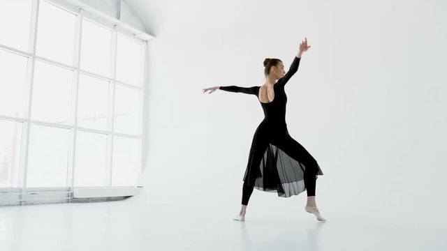 Attractive brunette ballerina in white pointe and black dress dancing in a white dance-hall near the big window 20s 4k