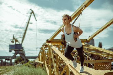 Naklejka premium Cheerful woman exercising with suspension trainer outdoor. Trx training against the background of metal parts.