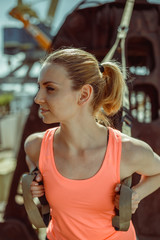 Close-up portrait of an athlete in uniform during exercise with TRX exercise outdoors. Beautiful young woman in an orange T-shirt.
