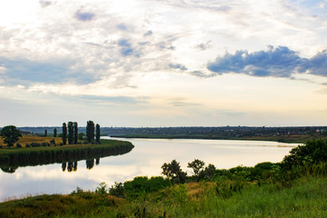 Summer evening dawn sky clouds reflected on a lake of water on the coast with green grass and forest trees