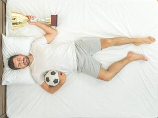 The sleeping sportsman hold a ball and a metal cup on the bed. view from above