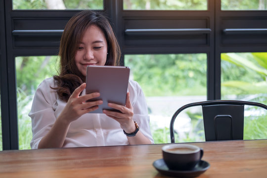 A beautiful Asian woman with smiley face holding and using tablet pc in modern cafe with green nature background