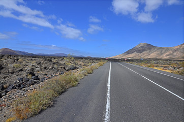 An empty road through volcanic landscape of Lanzarote, the Canary Islands. Volcanoes in the background and blue sky with picturesque clouds.