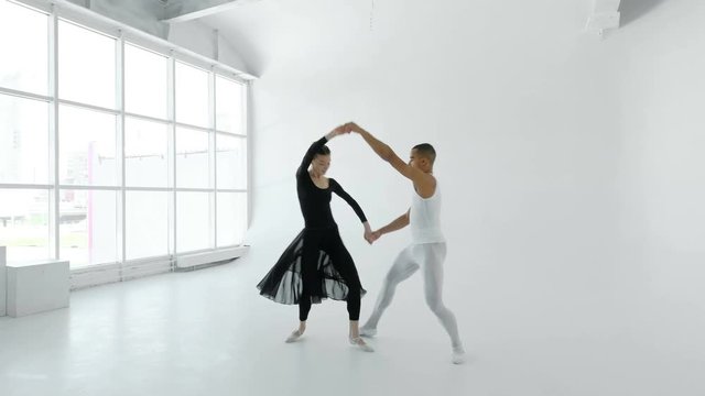 Young and attractive 20s ballet dancers in white and black gracefully dance in a white room with a large window 4k.