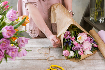 Female hands pack festive bouquet in wrapping paper. Unrecognizablel florist make floristry assemble with pink peony and wildflowers in workshop on wooden background. Woman at work