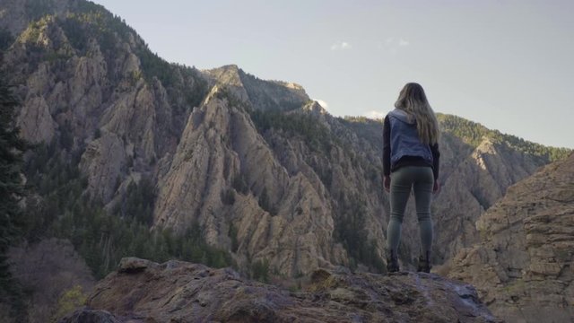 Adventurous Young Woman Stands (Back To Camera) Atop A Boulder And Looks Out At Mountain Vista 