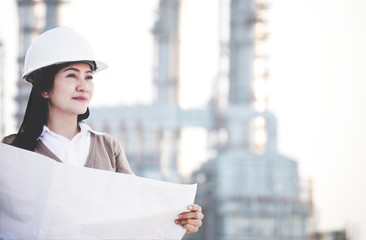 Engineer asia woman with hard hat holding blue print paper looking away inspecting progress at construction power plant site, safety control.  Engineer Concept.