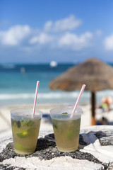 Two cocktails on the beach on a sunny day. Mojito drinks with straw. Umbrella and turquoise sea in the background out of focus.