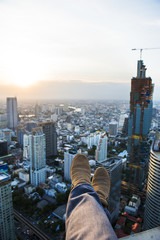 Legs hanging down from the roof top into the air with front aerial view over beautiful twilight of a big city in Bangkok, extreme activity, dangerous, relax.