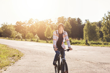 Cute teenage boy wearing shirt carrying backpack riding his bicycle having rest during his ride in suburb of city. Young schoolboy with trendy hairstyle resting outdoors with his modern bike