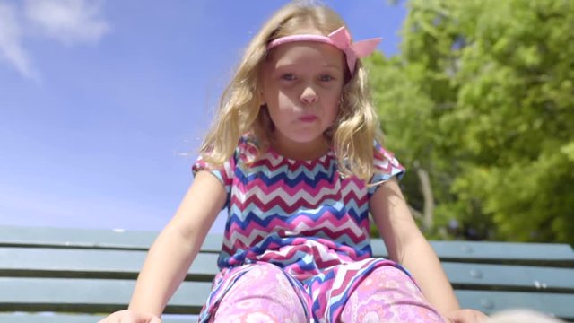 Cute Little Girl Swings Her Feet On Bench, She Holds Up Her Foot To Look At It, Gets Off Bench