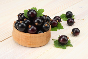 black currant in wooden bowl with green leaf on white wooden background.