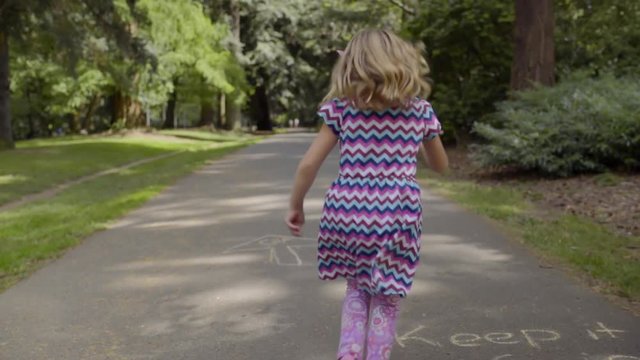 Little Girl Skips Down Path In Park (With Chalk Drawings On It) On Sunny Day 