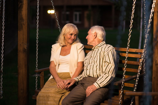 Couple Sitting On Porch Swing. Old Man And Woman, Evening.