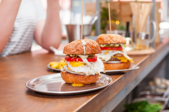 Two Tasty Beef Burgers with tomato, cheese and fried egg on a metal trays on the bar counter waiting to be served. Selective focus, close up. Fast food cafe cuisine. Space for text