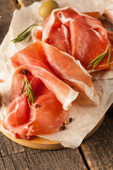 closeup of thin slices of prosciutto with mixed olives and paprika on wooden cutting board
