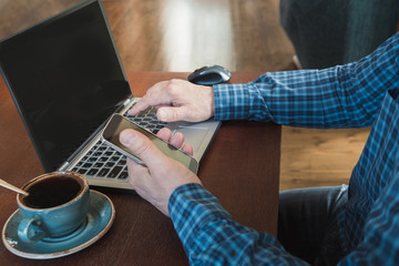 Side view shot of a man's hands using smart phone and laptop sitting at wooden table with cup of black coffee in interior.  Close up.