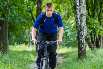 A seventeen-year-old boy, a teenager, rides a bicycle through the forest in the mountains. He is wearing a blue T-shirt and black sports pants, with a backpack behind his shoulders.