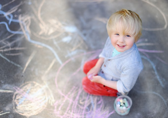 Naklejka premium Happy little kid boy sitting and drawing with colored chalk on asphalt