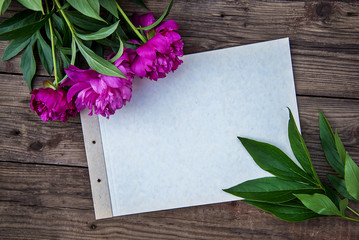 A sheet of paper and a few pink peonies on wooden background with copy space as a postcard