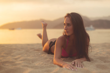 Attractive model with brown hair wearing bikini enjoying summer holidays lying on sandy beach at sunset sea and mountains in background