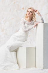 Fototapeta premium fashionable dress, beautiful blonde model, bride hairstyle and makeup concept - luxury young woman in white wedding gown sitting indoors on light background, female posing in the studio