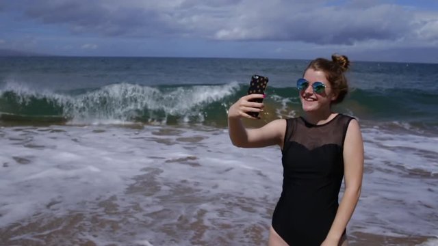 Slow Motion Shot Of Cute Teen Taking Fun Selfies On The Beach, Wave Crashes Behind Her, Maui, Hawaii