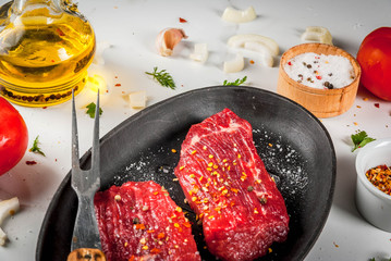 Meat, beef. Fresh raw steaks in a frying pan. Spices (salt, pepper), fresh vegetables - tomatoes, carrots, garlic, onions. On a white marble table, with a fork for meat and a knife.